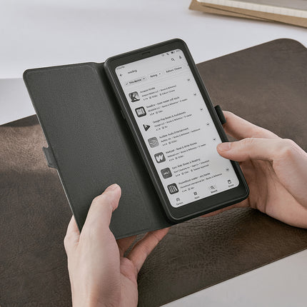 Hands holding an Onyx Boox Palma 2 in a dark flip cover, over a brown desk mat
